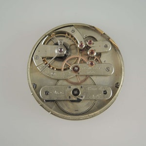 May include: Close-up of the inner workings of a silver pocket watch with a gold-toned winding mechanism. The watch face is visible with Roman numeral markings and the text "Swiss Made" is visible.
