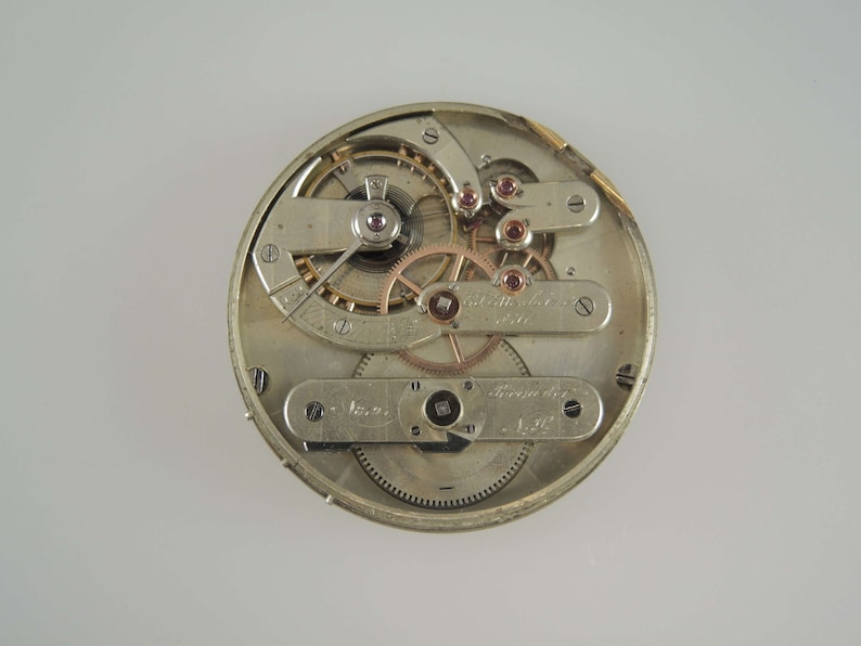 May include: Close-up of the inner workings of a silver pocket watch with a gold-toned winding mechanism. The watch face is visible with Roman numeral markings and the text "Swiss Made" is visible.