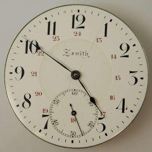 May include: Close-up of a vintage Zenith watch face. The white face features black Arabic numerals, a smaller seconds dial, and black hands. The brand name "Zenith" is printed in the center. The watch face has a metallic silver rim.