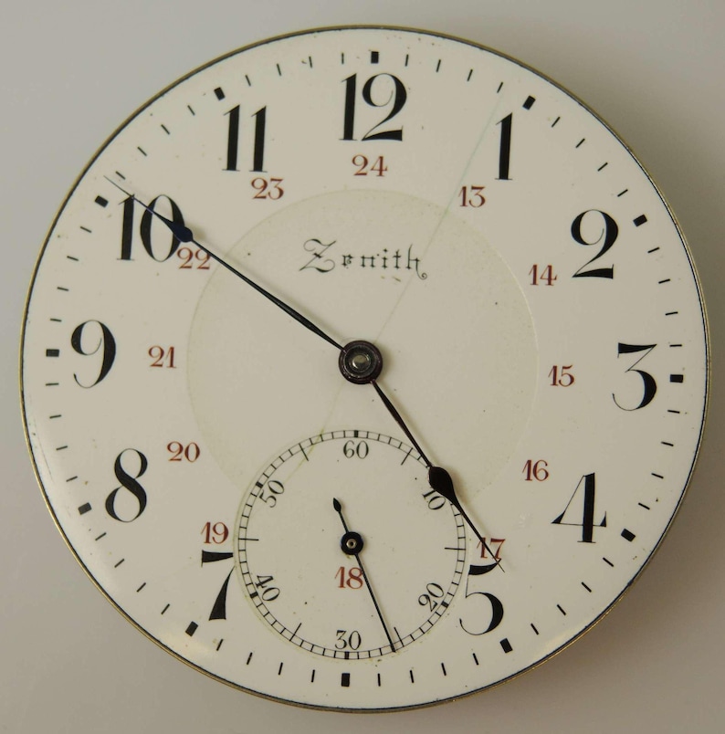 May include: Close-up of a vintage Zenith watch face. The white face features black Arabic numerals, a smaller seconds dial, and black hands. The brand name "Zenith" is printed in the center. The watch face has a metallic silver rim.