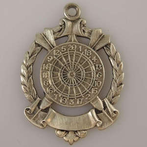 Könnte beinhalten: Silberfarbener Anhänger mit einer Dartscheibe mit den Zahlen 1-20, einem Lorbeerkranz und zwei gekreuzten Pfeilen mit einem Banner.