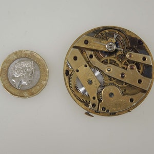 May include: Close-up of a gold-toned watch movement with intricate gears and a winding mechanism. The movement is exposed and visible, showing the inner workings of the watch.