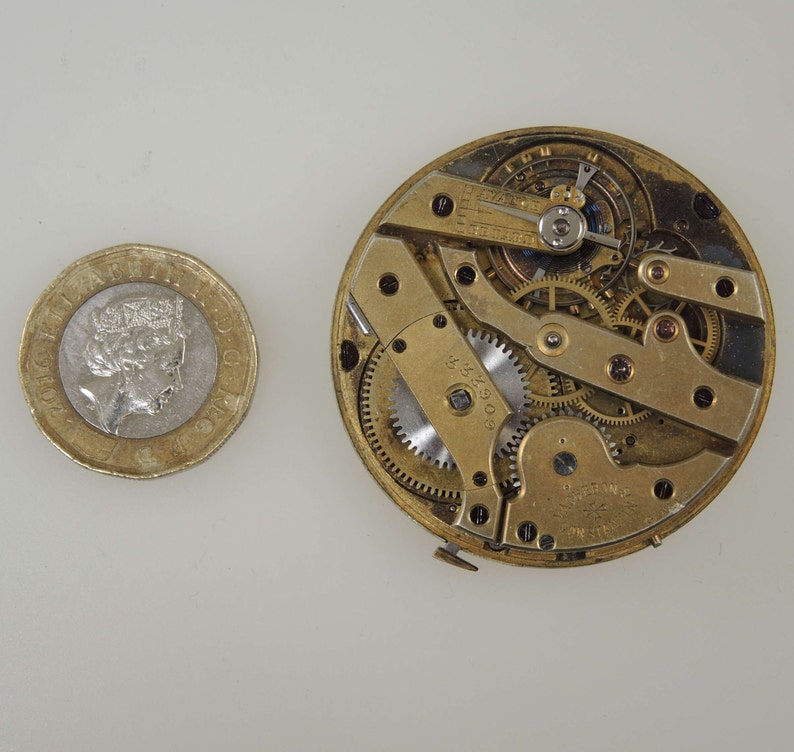 May include: Close-up of a gold-toned watch movement with intricate gears and a winding mechanism. The movement is exposed and visible, showing the inner workings of the watch.