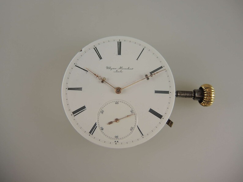Op de afbeelding: Een wit zakhorloge-wijzerplaat met zwarte Romeinse cijfers voor de uren en zwarte markeringen voor de minuten. De wijzerplaat heeft een goudkleurige opwindstift en wijzers. Op de wijzerplaat staat de tekst "Ulysse Nardin" en "Le Locle".