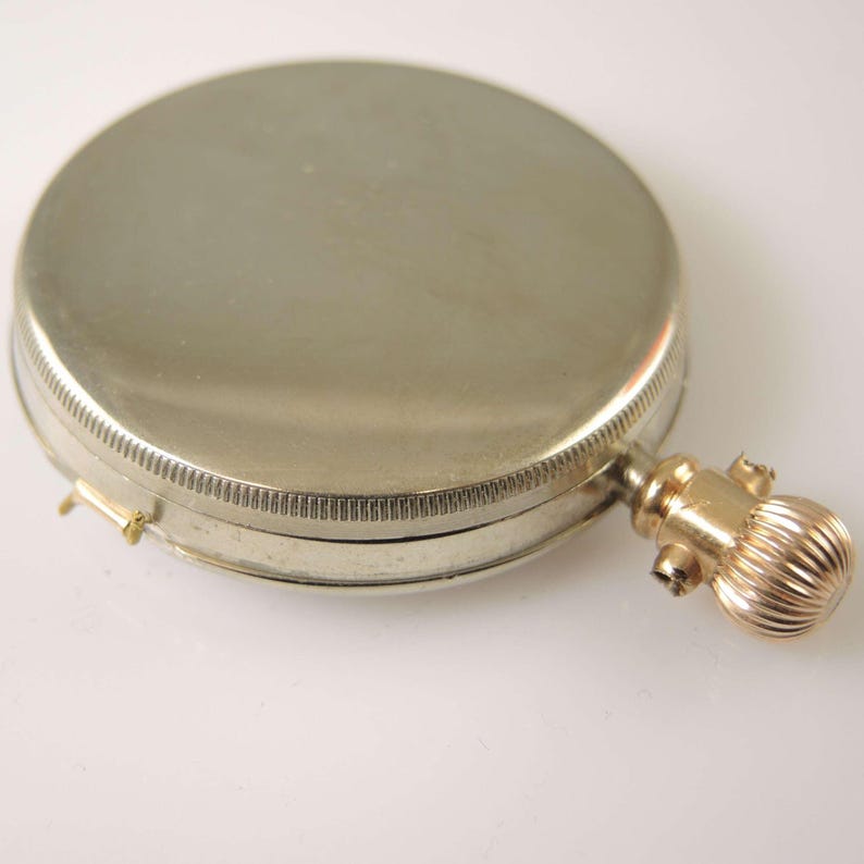 Puede incluir: Un estuche compacto redondo plateado con un borde acanalado y un asa dorada bulbosa. El estuche compacto est&aacute; cerrado y el asa tiene un dise&ntilde;o decorativo texturizado. La est&eacute;tica general es vintage.