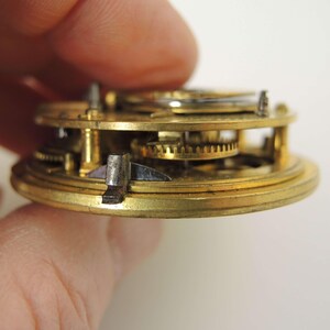 May include: Close-up of a vintage watch movement, showcasing intricate brass gears and components. The image highlights the mechanical details of the watch, with a focus on the precision and craftsmanship of the internal mechanisms. The watch movement is held by a person.