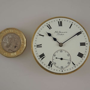 Op de afbeelding: Een vintage zakhorloge met een witte wijzerplaat, zwarte Romeinse cijfers en zwarte wijzers. Het horloge heeft een goudkleurige rand en de tekst "J.W. Benson London". Naast het horloge ligt een Britse twee-pond munt.