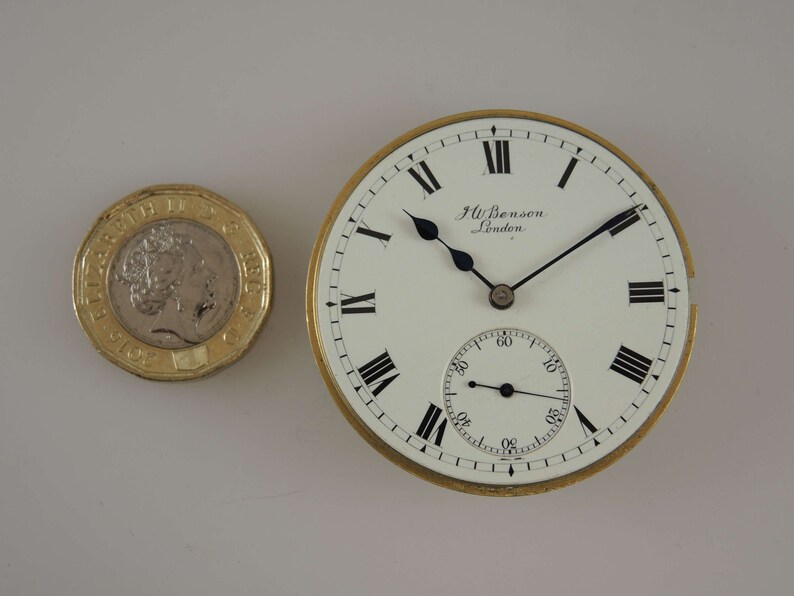 Op de afbeelding: Een vintage zakhorloge met een witte wijzerplaat, zwarte Romeinse cijfers en zwarte wijzers. Het horloge heeft een goudkleurige rand en de tekst "J.W. Benson London". Naast het horloge ligt een Britse twee-pond munt.