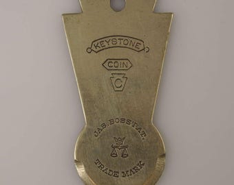 Puede incluir: Una llave en blanco de Keystone Coin de latón con el texto "KEYSTONE COIN" y "JAS. BOSSSTAT. TRADE MARK" con un diseño de corona y balanza.