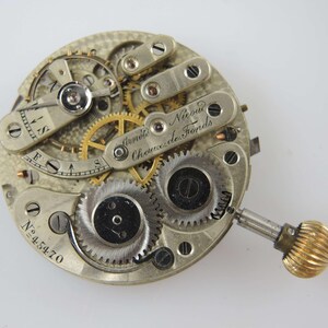 May include: Close-up of a vintage watch movement with intricate gears, screws, and a winding mechanism. The watch mechanism has a silver finish with gold-colored gears and a gold-colored winding crown. The text "Arnold Nicoud Chaux de Fonds" is visible.