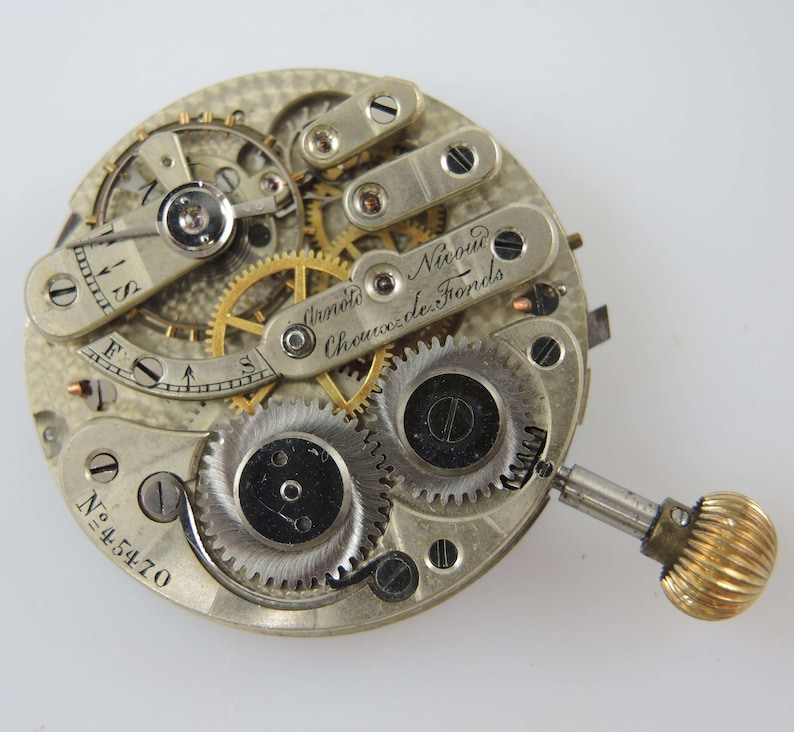 May include: Close-up of a vintage watch movement with intricate gears, screws, and a winding mechanism. The watch mechanism has a silver finish with gold-colored gears and a gold-colored winding crown. The text "Arnold Nicoud Chaux de Fonds" is visible.
