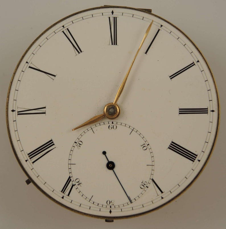 May include: A white pocket watch face with black Roman numeral hour markers and black minute markers. The watch has gold hands and a small seconds dial at the bottom.