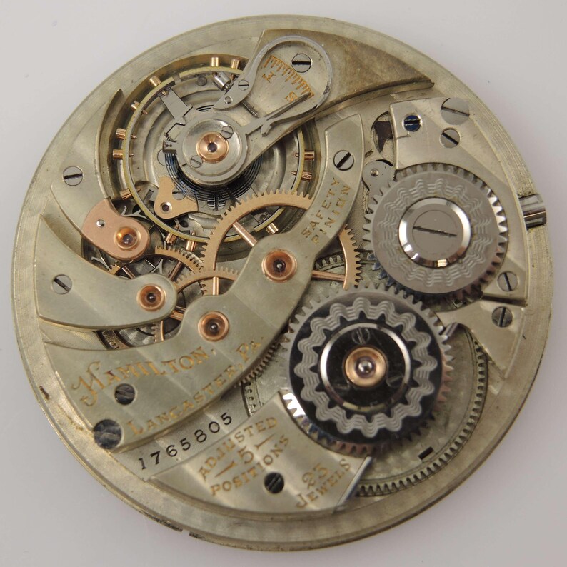 May include: Close-up of a vintage watch movement. The intricate mechanism features numerous gears, springs, and levers in silver and gold tones. The watch face includes the text "Hamilton Lancaster Pa." and "25 Jewels".