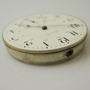 May include: Close-up of a vintage watch face with a white background. The watch face has black and red numerals, with the number 12 prominently displayed. The watch casing is silver and shows signs of wear.