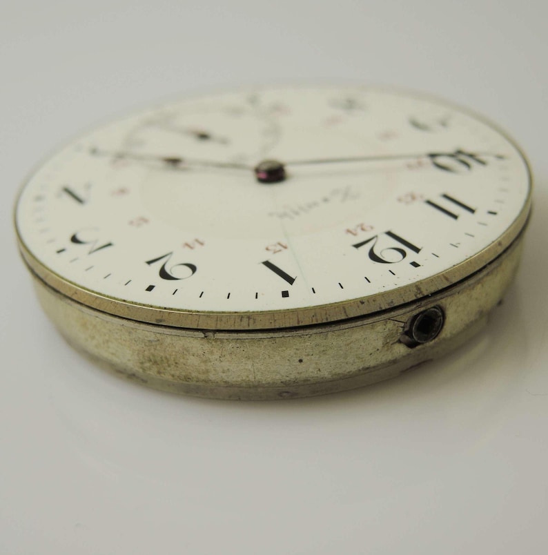 May include: Close-up of a vintage watch face with a white background. The watch face has black and red numerals, with the number 12 prominently displayed. The watch casing is silver and shows signs of wear.