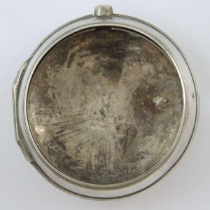 Könnte beinhalten: Ein offenes, silberfarbenes Taschenuhrgehäuse. Das Innere weist Gebrauchsspuren und Alter auf, mit einer strukturierten, angelaufenen Oberfläche. Das Gehäuse hat eine kreisförmige Form mit einer kleinen Schlaufe oben und einem dekorativen Rand.
