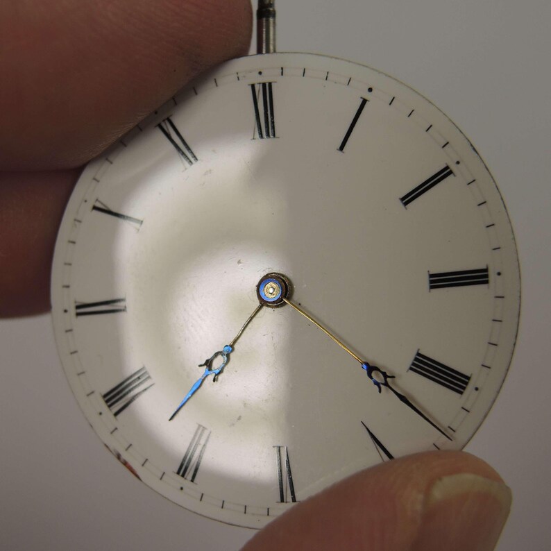 May include: A vintage watch face with a white background and black Roman numerals. The watch hands are blue and gold. The watch face is held between two fingers.