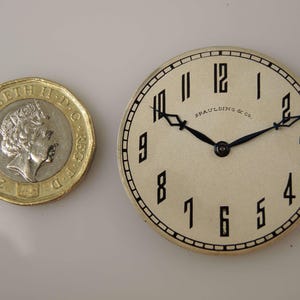 May include: A white watch face with black Roman numeral hour markers and black hands. The watch face is surrounded by a silver-coloured metal case. The text "Spaulding & Co." is printed on the watch face.
