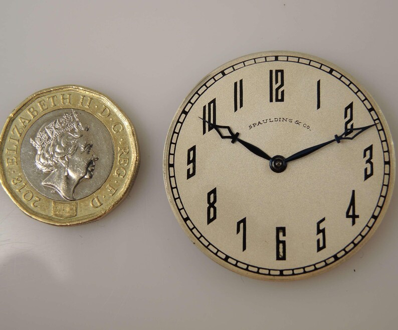 May include: A white watch face with black Roman numeral hour markers and black hands. The watch face is surrounded by a silver-coloured metal case. The text "Spaulding & Co." is printed on the watch face.