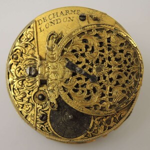 May include: A gold pocket watch with intricate engravings and a delicate openwork design. The watch face is visible and features the inscription "De Charme London".