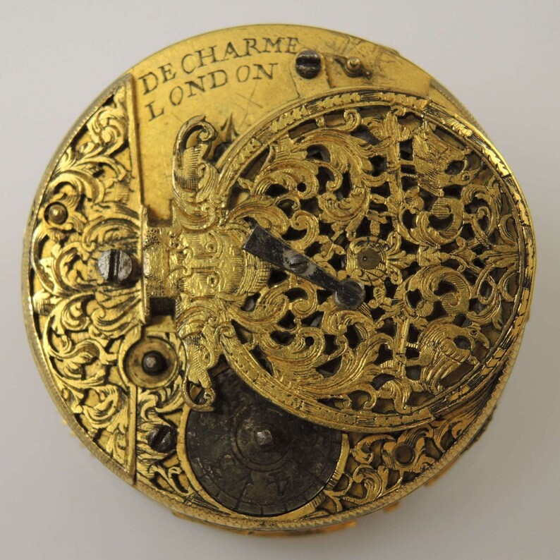 May include: A gold pocket watch with intricate engravings and a delicate openwork design. The watch face is visible and features the inscription "De Charme London".