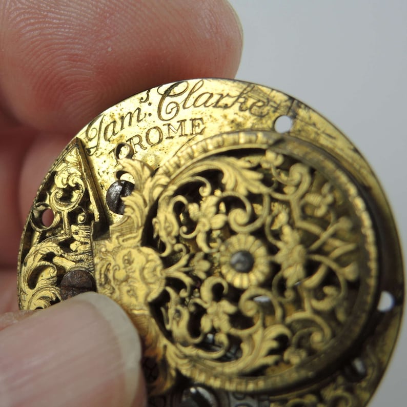 May include: A gold-colored pocket watch face with intricate floral designs and the inscription "Jam: Clarke ROME".