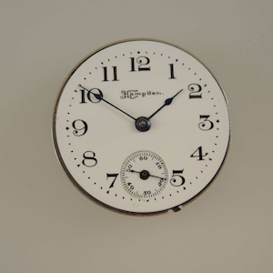 Puede incluir: Una esfera de reloj blanca con agujas negras y marcadores de hora en n&uacute;meros romanos negros. La esfera del reloj tiene el texto "The Hampden." impreso en ella.