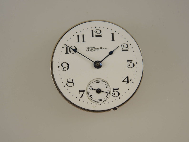 Puede incluir: Una esfera de reloj blanca con agujas negras y marcadores de hora en n&uacute;meros romanos negros. La esfera del reloj tiene el texto "The Hampden." impreso en ella.