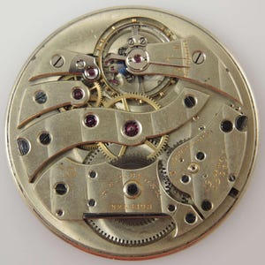 May include: Close-up view of a silver pocket watch movement with intricate gears and a gold-coloured winding stem. The watch is labelled "B.W. Raymond" and "15 Jewels."