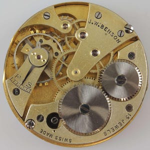 Puede incluir: Primer plano de un movimiento de reloj de bolsillo dorado con el texto "J.W. BENSON" y "15 JEWELS" visible. El movimiento est&aacute; etiquetado como "SWISS MADE".