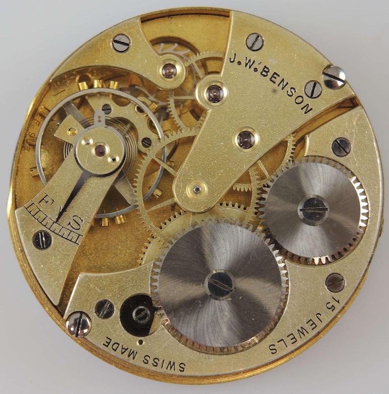 Puede incluir: Primer plano de un movimiento de reloj de bolsillo dorado con el texto "J.W. BENSON" y "15 JEWELS" visible. El movimiento est&aacute; etiquetado como "SWISS MADE".