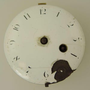 Puede incluir: Una esfera de reloj blanca antigua con n&uacute;meros negros. La esfera est&aacute; agrietada y da&ntilde;ada, con una secci&oacute;n faltante cerca del n&uacute;mero seis. Hay dos agujeros de color marr&oacute;n oscuro en el centro.