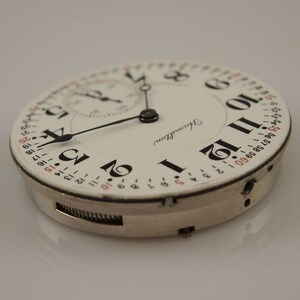May include: A white pocket watch with black Roman numeral hour markers and black hands. The watch face also has a smaller dial with black hands and red numbers. The watch is labeled "Hamilton".