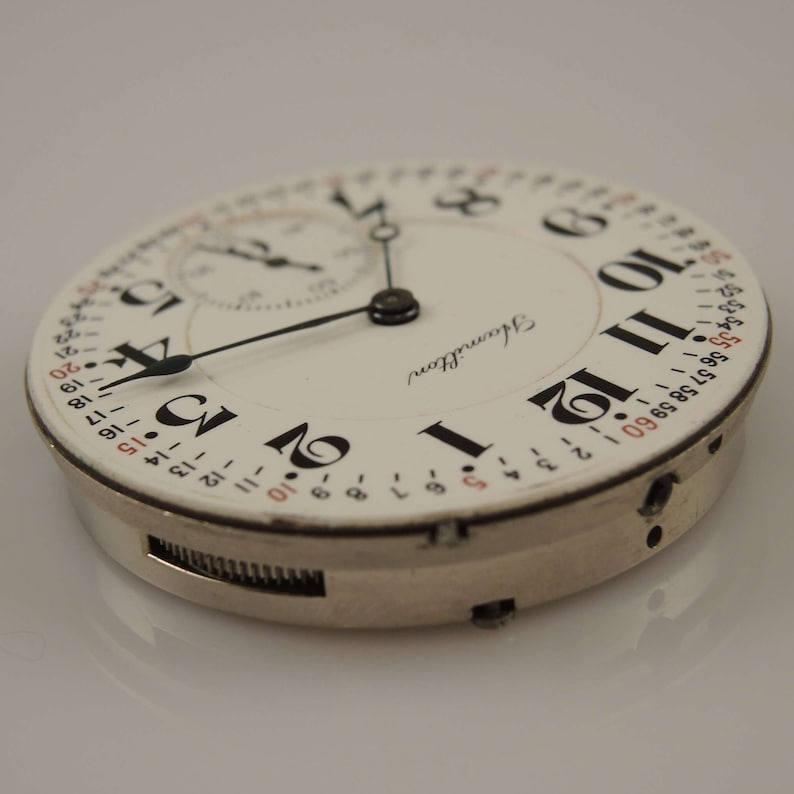 May include: A white pocket watch with black Roman numeral hour markers and black hands. The watch face also has a smaller dial with black hands and red numbers. The watch is labeled "Hamilton".