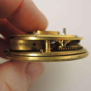 May include: Close-up of a vintage pocket watch mechanism. The brass components include gears, a balance wheel, and other intricate parts. The watch is held by a hand, showcasing the detailed craftsmanship of the antique timepiece.