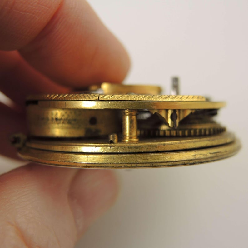 May include: Close-up of a vintage pocket watch mechanism. The brass components include gears, a balance wheel, and other intricate parts. The watch is held by a hand, showcasing the detailed craftsmanship of the antique timepiece.