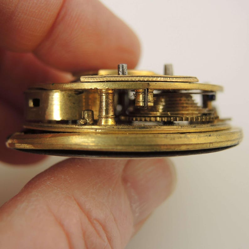 May include: Close-up of a vintage watch movement, showcasing intricate brass gears and components. The mechanism is held in a hand, revealing the detailed craftsmanship of the timepiece. The golden color of the brass contrasts with the darker background, highlighting the precision of the watch's internal workings.