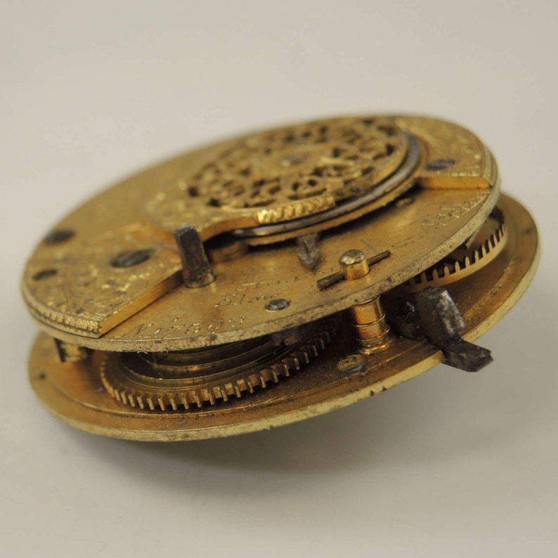 May include: Close-up of a gold-toned pocket watch movement with intricate gears and a decorative design. The watch is disassembled and shows the inner workings.
