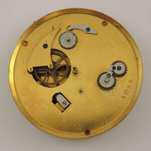 May include: Close-up of the inner workings of a gold-toned pocket watch. The watch face is missing, but the intricate gears and levers are visible. The number "10592" is printed on the watch case.