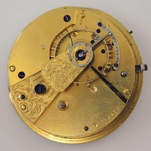May include: A detailed close-up of a gold-coloured watch movement, showcasing intricate gears, screws, and a balance wheel. The movement has engraved floral patterns and a serial number. The hands are black, and the design is ornate and detailed.