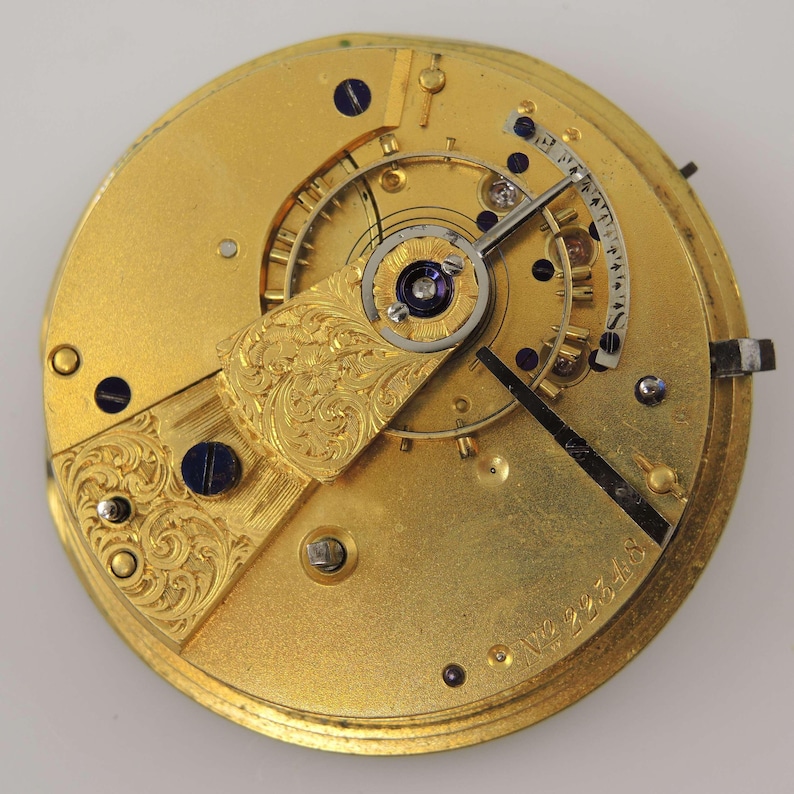 May include: A detailed close-up of a gold-coloured watch movement, showcasing intricate gears, screws, and a balance wheel. The movement has engraved floral patterns and a serial number. The hands are black, and the design is ornate and detailed.
