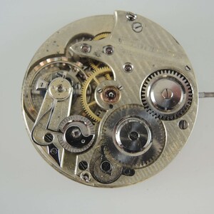 May include: Close-up of a silver pocket watch movement with intricate gears and a winding stem. The watch face is not visible.