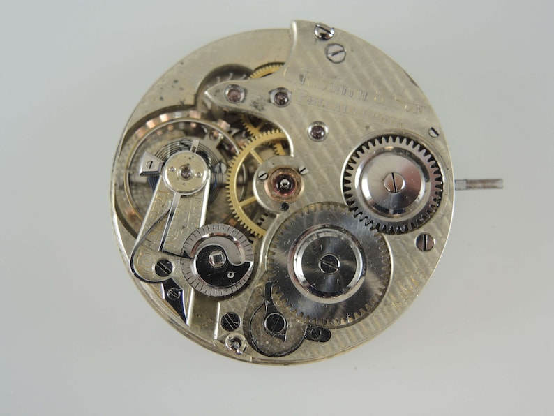 May include: Close-up of a silver pocket watch movement with intricate gears and a winding stem. The watch face is not visible.
