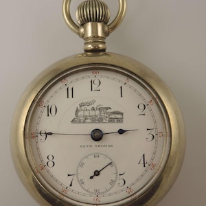 Peut inclure: Une montre de poche Seth Thomas argentée avec un cadran blanc. Le cadran comporte des chiffres noirs, un petit cadran des secondes et une illustration détaillée d'un train. La montre a une tige de remontoir et une boucle pour une chaîne.