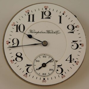 K&ouml;nnte beinhalten: Eine wei&szlig;e Taschenuhr mit schwarzen r&ouml;mischen Ziffern f&uuml;r die Stunden und schwarzen Zeigern. Das Zifferblatt der Uhr hat einen kleineren, konzentrischen Kreis mit schwarzen arabischen Ziffern f&uuml;r die Minuten und schwarzen Zeigern. Das Zifferblatt der Uhr tr&auml;gt auch den Text "Hampden Watch Co." in Schwarz.