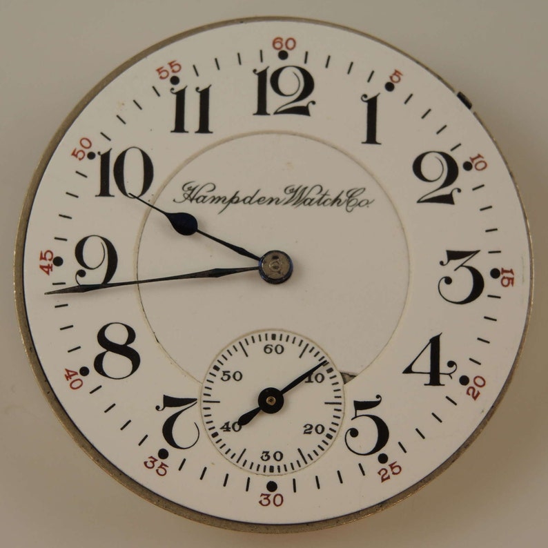K&ouml;nnte beinhalten: Eine wei&szlig;e Taschenuhr mit schwarzen r&ouml;mischen Ziffern f&uuml;r die Stunden und schwarzen Zeigern. Das Zifferblatt der Uhr hat einen kleineren, konzentrischen Kreis mit schwarzen arabischen Ziffern f&uuml;r die Minuten und schwarzen Zeigern. Das Zifferblatt der Uhr tr&auml;gt auch den Text "Hampden Watch Co." in Schwarz.
