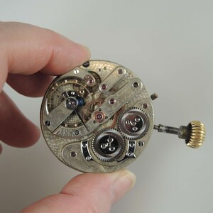 May include: A silver pocket watch movement with intricate gears and a gold winding stem.