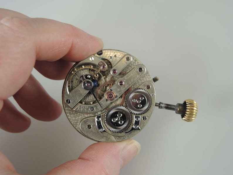 May include: A silver pocket watch movement with intricate gears and a gold winding stem.