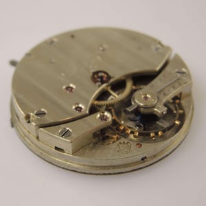 May include: Close-up of a vintage watch movement, showcasing intricate gears, screws, and metal components. The watch mechanism is silver-toned with small ruby jewels and a detailed design, highlighting the craftsmanship of the timepiece.