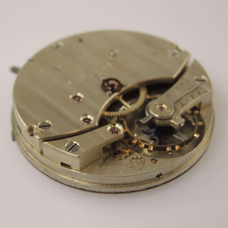 May include: Close-up of a vintage watch movement, showcasing intricate gears, screws, and metal components. The watch mechanism is silver-toned with small ruby jewels and a detailed design, highlighting the craftsmanship of the timepiece.